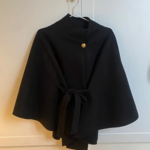 Svart cape med knytband - Snygg svart cape med knytband och en dekorativ knapp framtill. Perfekt för att ge en elegant touch till din outfit. Den har en lös passform och är lätt att bära över andra plagg. Passar en storlek S-M. Jag är 174
