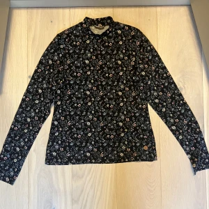 Svart blommig långärmad tröja från Kappahl  - Säljer en svart långärmad tröja med ett fint blommigt mönster i ljusa färger. Tröjan är tillverkad i bomull med lite elastan för extra komfort. Perfekt för en stilren och bekväm look.
