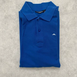 Blå pikétröja från J.Lindeberg - Säljer en stilren blå pikétröja från J.Lindeberg. Tröjan har en klassisk krage och knappar framtill. Perfekt för en avslappnad men ändå snygg look. Sparsam använt 