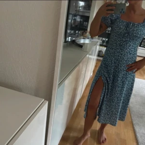 Blommig blå klänning med slits - Säljer en söt blå klänning med vita blommor och en smickrande slits. Klänningen har korta ärmar och en fin passform som passar perfekt för sommardagar. Perfekt för en avslappnad och stilren look. Aldrig använd 