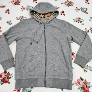 Grå huvtröja från Burberry (storlek L men passar M också)  - Säljer en snygg grå huvtröja från Burberry med klassiskt rutigt foder. Tröjan har dragkedja och snörning i huvan. Perfekt för en stilren look.