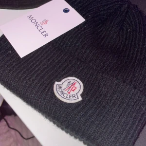 Moncler mössa - Snygg svart mössa från Moncler med ribbad design och broderad logotyp framtill. Perfekt för att hålla värmen med stil under kyliga dagar. Den är i topp skick och nästan aldrig använd!