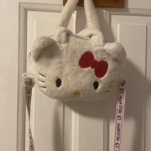 Vit Hello Kitty axelväska - Söt och fluffig vit axelväska formad som Hello Kittys ansikte med en röd rosett. Väskan har ett mjukt material och ett justerbart axelband med Hello Kitty-logga. Perfekt för att lägga till en gullig touch till din outfit!
