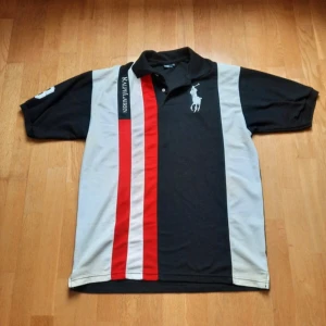 Svart och grå pikétröja från Ralph Lauren - Säljer en snygg pikétröja från Ralph Lauren i svart och grått med röda och vita ränder. Tröjan har korta ärmar och en klassisk krage. Den ikoniska loggan är broderad på bröstet. Perfekt för en stilren look.