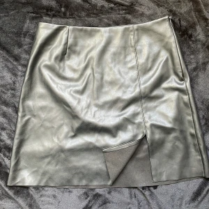 Svart skinnkjol från Vero moda - Snygg svart skinnkjol från Vero moda med en liten slits framtill. Kjolen har en dragkedja på sidan för enkel på- och avtagning. Längden är ungefär till mitten av låren, lite längre och är nögmidjad. Skriv för fler bilder/frågor!🫶🏻