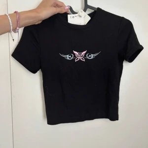 Svart croppad t-shirt med fjärilsbroderi BERSHKA Y2K - Säljer en svart croppad t-shirt med ett snyggt broderi av en rosa fjäril och blå detaljer på bröstet. Perfekt för en trendig look. Kortärmad och i mjukt material. Ny med prislapp!