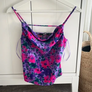 Blommig linnetopp från BikBok - Säljer en färgglad blommig linnetopp från Bibok. Toppen har tunna axelband och ett härligt mönster i lila, rosa och grönt. Lite tyg hänger fram över bröstet så den ger en festlig känsla!