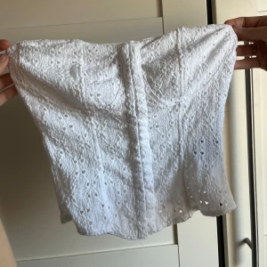 Vit spets korsett - Säljer en söt vit korsett med spetsdetaljer. Den är axelbandslös och har en vacker broderad design. Perfekt för att ge en romantisk touch till din outfit.