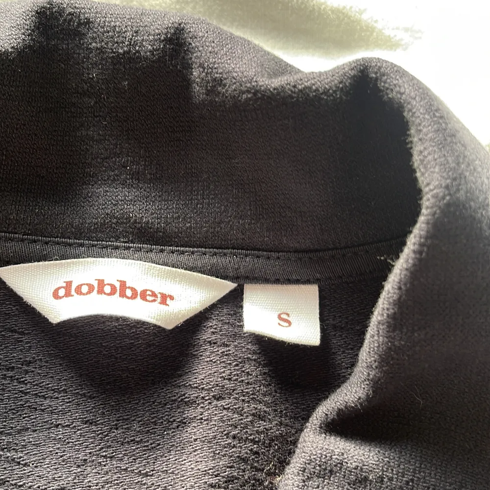 Snygg svart långärmad pikétröja från Dobber med dragkedja vid halsen. Perfekt för en stilren och avslappnad look. Tröjan har en klassisk krage och är tillverkad i ett bekvämt material.. Neuletakit & Villapaidat.
