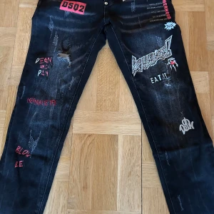 Svarta jeansbyxor från Dsquared2 - Unika svarta jeansbyxor från Dsquared2 med coola tryck och slitningar. Byxorna har en edgy design med text och grafiska element i vitt och rött. Perfekta för dig som vill sticka ut med en trendig stil.