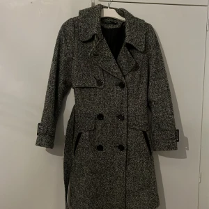 Grå kappa från KappAhl - Snygg grå kappa från KappAhl med dubbelknäppning och klassisk krage. Den har långa ärmar och ett bälte i midjan för en elegant siluett. Perfekt för kyligare dagar.