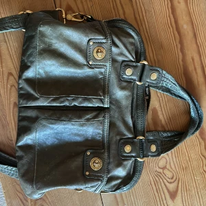 Grön skinnväska från Marc Jacobs - Snygg svart skinnväska från Marc Jacobs med dragkedja och guldfärgade detaljer. Väskan har två framfickor med vridlås och ett mönstrat innerfoder med logotyp. Perfekt för att bära dina dagliga nödvändigheter med stil. Vintage och väldigt snygg