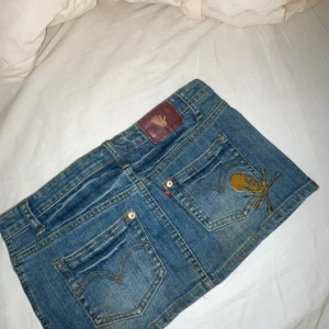 Jeanskjol med broderad detalj - Snygg jeanskjol med en cool broderad dödskalle på bakfickan. Kjolen har en klassisk femficksdesign och slitna detaljer framtill för en trendig look. Perfekt för en avslappnad stil. Midjemått rakt över är 38cm💖INTRESSEKOLL!