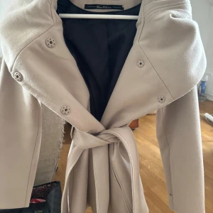 Beige kappa från new yorker - Elegant beige kappa från new torkar  med knappar och ett bälte i midjan. Har även en luva som går ner lite över axlarna. Mycket bra skick skriv för mer info 