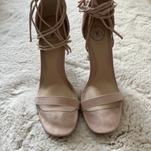 Beige klackskor med snörning - Snygga beige klackskor med öppen tå och snörning runt ankeln. Perfekta för en elegant look. Tillverkade i mocka med en smal klack som ger en stilren siluett.