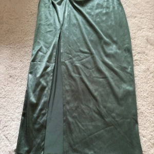 Grön långkjol från ASOS - Grön långkjol från ASOS med en elegant slits och en dekorativ ringdetalj i midjan. Kjolen har en glansig finish som ger en lyxig känsla. 