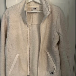 Vit teddyjacka från The North Face - Mysig vit teddyjacka från The North Face med dragkedja och två fickor framtill. Perfekt för kyliga dagar när du vill hålla dig varm och bekväm. Jackan har en normal passform och är långärmad.