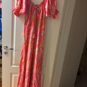 Färgglad långklänning från Coconutmilk By Stajl - Säljer en färgglad långklänning i rosa och orange från Coconutmilk By Stajl. Klänningen har ett tie-dye mönster och offshoulder design med volangdetaljer. Perfekt för sommarens varma dagar!