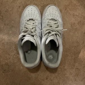 Vita Nike Air Force 1 - Säljer ett par klassiska vita Nike Air Force sneakers. De har en stilren design med snörning och perforerade detaljer på ovansidan för ventilation. Perfekta för en casual look.