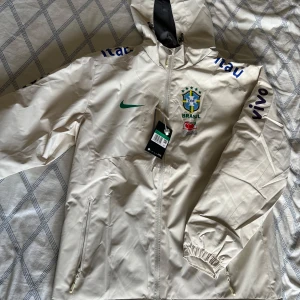 Vit Nike Brazil Jacka  - Säljer en vit Brasilien jacka från Nike med huva och dragkedja. Jackan har broderad logga och text på bröstet och ärmarna. Insidan är fodrad med mjukt material för extra komfort, och en innerficka. Perfekt för vårdagar med varierande väder. Accepterar lämpliga erbjudanden. Han är liten i storleken, står XL på etikett men passar definitivt en S/M. Meddela mig vid eventuella mått. Pris kan diskuteras vid snabb affär. 