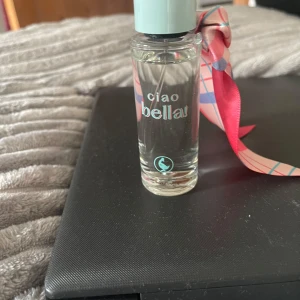 Ciao Bella parfym - Charmig parfymflaska med texten 'ciao bella' i en stilren design. Flaskan är genomskinlig med ett ljusblått lock och dekorerad med ett färgglatt band i rosa och blått. Perfekt för den som vill ha en fräsch och ungdomlig doft.