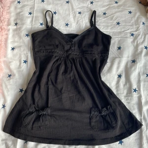 Svart ärmlös topp med knappar - Säljer en svart ärmlös topp med smala axelband och knappar framtill. Toppen har en v-ringad design och små fickor med rynkdetaljer. Perfekt för en stilren look.