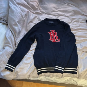 Svart tröja från Bondelid - Säljer en mörkblå tröja från Bondelid med röd logotyp på bröstet. Tröjan har långa ärmar och ribbade muddar med vita ränder vid ärmslut och nederkant. Perfekt för en stilren look. Passar st. S också.