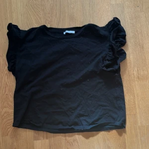 Svart topp från Zara - Säljer en svart topp från Zara med volangdetaljer på ärmarna. Toppen har en rund halsringning och är kortärmad. Perfekt för en stilren look.
