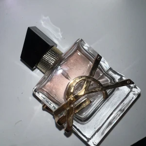 Parfym från Yves Saint Laurent - Elegant parfymflaska med en unik design från Yves Saint Laurent. Flaskan är genomskinlig med en ljusrosa vätska och har en svart kork. Den pryds av ett guldigt metallband som ger en lyxig känsla.