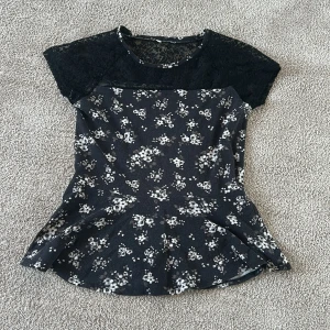 Svart blommig topp/tunika med spetsdetaljer - Snygg svart topp/tunika med vita blommor och spetsdetaljer på axlarna. Står ej storlek men jag skulle säga att den passar ungefär 11-17 åringar🫰🏼🫰🏼