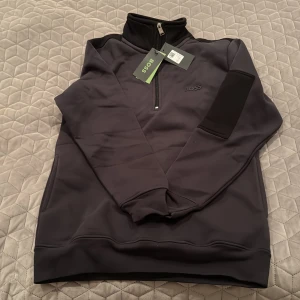 Svart tröja med dragkedja från Boss - Säljer en stilren svart tröja från Boss med dragkedja och hög krage. Perfekt för en avslappnad men snygg look. Tröjan har långa ärmar och en diskret logga på bröstet.