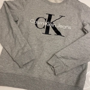 Grå sweatshirt från Calvin Klein Jeans - Säljer en stilren grå sweatshirt från Calvin Klein Jeans med det ikoniska CK-logotypen tryckt på framsidan. Tröjan har långa ärmar och en rund halsringning. Perfekt för en avslappnad look.