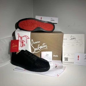Svarta sneakers med nitar från Christian Louboutin - Snygga svarta sneakers från Christian Louboutin i mocka med ikoniska röda sulor. Skorna har svarta nitar på tån och snörning för en edgy look. Perfekta för att ge din outfit en lyxig touch, helt nya 