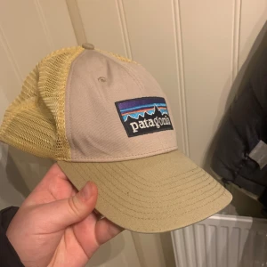 Beige keps från Patagonia - Snygg beige keps från Patagonia med mesh-bakdel för extra ventilation. Framsidan har en broderad logotyp i färgglada toner. Perfekt för sommarens äventyr! Legat i en garderob ett bra tag men är i bra skick (nypris 450kr) ( mitt pris 170kr+ 29kr frakt)