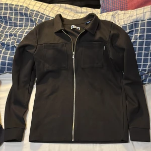 Svart overshirt/kofta från Jack & Jones - Säljer en stilren svart overshirt från Jack & Jones med dragkedja framtill. Jackan har två bröstfickor och är perfekt för en casual look. Den är i storlek 176 men lite lång i modellen så passar dig runt ca 180 cm. Aldrig använd utan har bara legat i garderoben. Pris kan diskuteras vid snabb affär