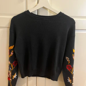 Svart tröja med broderade ärmar från H&M Divided - Säljer en svart tröja från H&M Divided med färgglada broderier på ärmarna. Tröjan har en rund halsringning och långa ärmar. Perfekt för att ge en färgklick till din outfit!