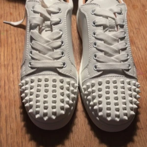 Vita christian louboutin  - Snygga vita sneakers med nitar på tån och klassisk röd sula. Skorna har snörning och är tillverkade i skinn. Perfekta för att ge din outfit en edgy touch. Storlek 38.