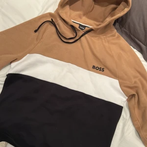 Beige, vit och svart hoodie från Boss - Säljer en snygg hoodie från Boss i beige, vit och svart. Den har en klassisk design med huva och dragsko. Perfekt för en avslappnad stil.