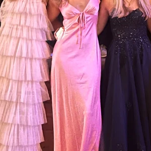 Balklänning - Säljer en elegant rosa långklänning med knyt framtill och baktill. Klänningen har en v-ringad design och är perfekt för speciella tillfällen. Materialet ser ut att vara mjukt och följsamt, vilket ger en fin passform. Använd en gång