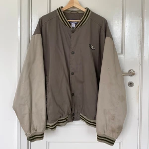 grön & beige collegejacka från B.U.M - snygg coch bekväm ollegejacka i gröna och beigea toner från B.U.M equipment i 100% bomull. jackan har rymliga ärmar, ribbade muddar med gröna ränder och tryckknappar framtill. klassisk college-look med logga på bröstet och skön oversized passform!