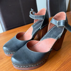 Svarta träklackar från Ten Points - Chunky träklackar från Ten Points i svart skinn med bred klack och justerbart spänne runt vristen. Snygg kontrastsöm och klassisk rund tå. Insidan är fodrad i ljusrosa. Perfekta för dig som gillar retrovibbar och vill sticka ut med en cool och stabil klack. Endast använda en gång. 