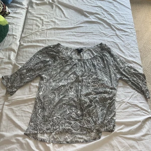 Svartvit blus med paisleymönster - Säljer en luftig svartvit blus från Lindex med paisleyinspirerat mönster. Blusen har trekvartsärm och en bred resår vid halsen som ger en avslappnad vibe. Perfekt att styla med jeans eller kjol för en cool look.