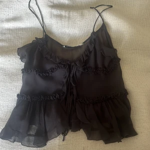 Svart volangig linnetopp från Zara - Supersöt svart linnetopp från Zara med tunna axelband, volanger och snygga rynkade detaljer. Toppen har knyt framtill och är gjord i ett lätt, transparent tyg som ger en luftig känsla. Knappt använd och känns som ny, pris går att diskuteras