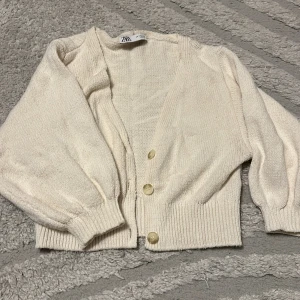 Beige kofta från Zara med knappar - Superfin beige kofta från Zara med tre knappar framtill och ribbade muddar. Koftan har en croppad passform och ballongärmar som ger en trendig look. Perfekt att slänga över en topp för en chill stil.