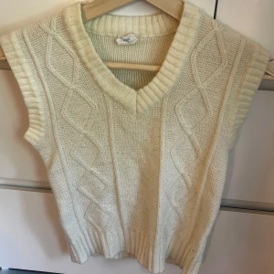 Vit stickad väst med v-ringning - Säljer en krämvit stickad väst med v-ringning och snyggt kabelstickat mönster framtill. Västen har breda ribbade kanter vid hals och nederkant, vilket ger en klassisk och trendig look. Perfekt att styla över en skjorta eller t-shirt.