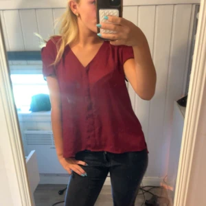 Vinröd blus med v-ringning från H&M - Säljer en vinröd blus från H&M i storlek 36. Blusen har v-ringning, korta ärmar och knappar framtill. Den är lätt och flowy, perfekt till jeans eller kjol. Enkel och stilren design som passar till många olika looks.