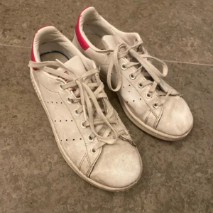 Adidas Stan Smith vita sneakers med rosa detalj - Klassiska Adidas Stan Smith sneakers i vitt skinn med rosa hälkappa och perforerade ränder på sidorna. Skorna har snörning och platt sula, samt rund tå. Perfekta för en clean och sportig look.💕💕