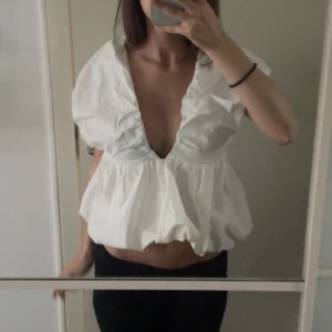 Vit blus med djup v-ringning och volang - Supersöt vit blus med djup v-ringning och volangkant runt halsen. Puffiga korta ärmar och knytning i ryggen ger en trendig look. Blusen är luftig och har en lätt croppad passform, perfekt för att sticka ut.