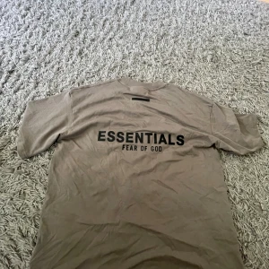 Beige Essentials Fear of God t-shirt - Snygg beige t-shirt från Essentials Fear of God med svart logga på ryggen. Klassisk rund hals och korta ärmar. Tillverkad i mjuk bomull som känns skön mot huden. Snygg och i helt nytt perfekt för dig som klär dig fräscht med fräscha färger 