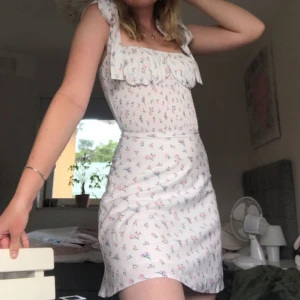 Vit blommig klänning  - Supersöt vit kortklänning med små blommor i rosa och blått. Klänningen har smockad överdel och knytband på axlarna. 